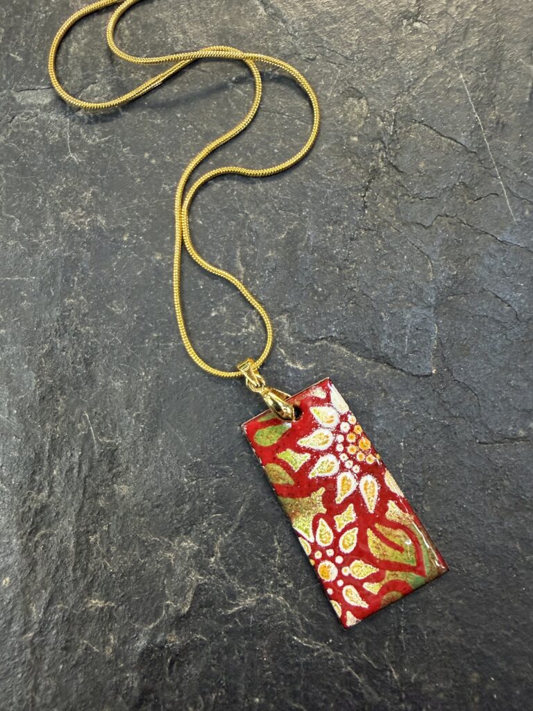 A red, white, green and yellow enamelled pendant on a gold chain with a daisy design made at Rainbow Glass Studios on the beginners course